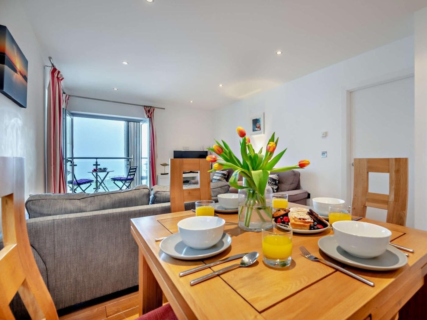 A dining area with a table set for breakfast, a vase of tulips, and a balcony with a view.