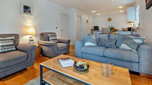Ein modernes Wohnzimmer mit einer Couch, zwei Sesseln, einem Couchtisch mit Obstschale und einer Küche im Hintergrund.