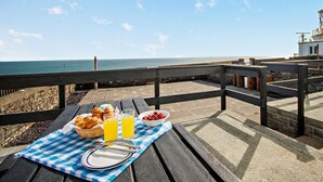 Un pique-nique installé sur une terrasse en bord de mer, avec une nappe à carreaux, des croissants, des fruits et du jus d’orange.