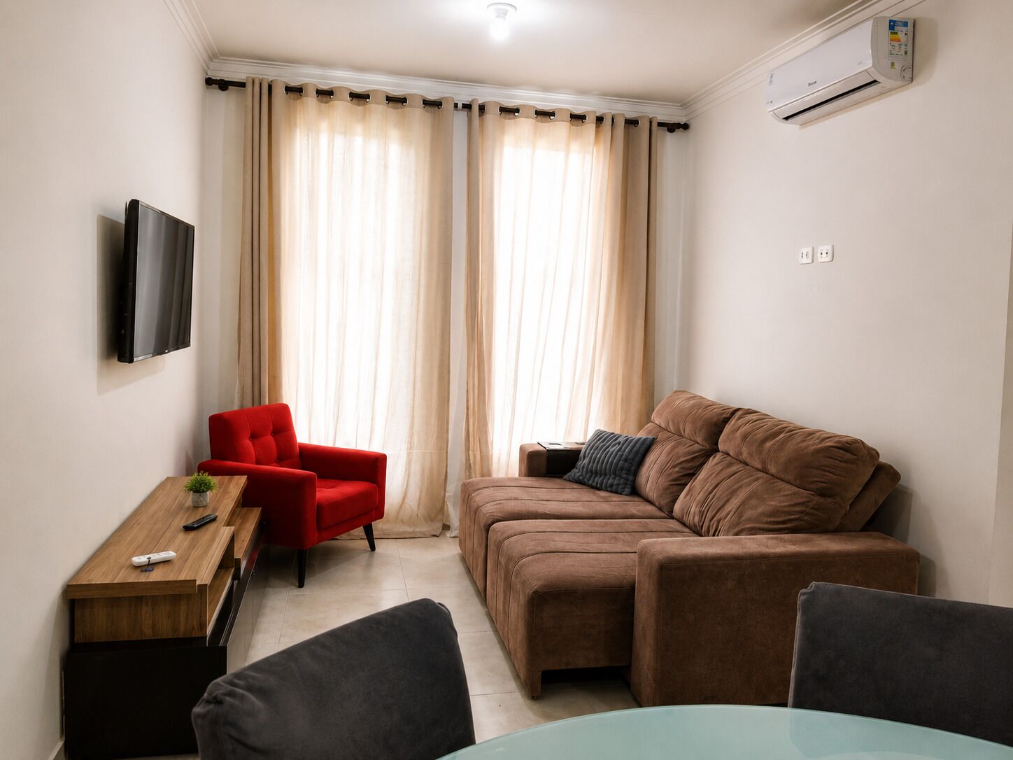 A modern living room with a brown sofa, a red armchair, a wooden side table, and a flat-screen TV mounted on the wall.