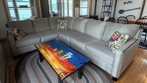 Ein Wohnzimmer mit einer grauen Eckcouch, einem bunten Couchtisch, einem gewebten Teppich und Blick auf einen See durch große Fenster.