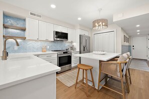 A modern kitchen with white cabinetry, stainless steel appliances, a herringbone tile backsplash, and a central island with stools. - Ocean Side Condo | Steps to the Beach (Ocean City)