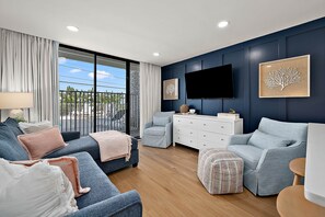 A modern living room with a sofa, armchairs, a TV, and a view through large windows. - Ocean Side Condo | Steps to the Beach (Ocean City)
