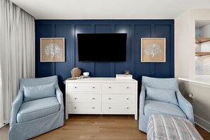 A room with a dark blue wall, a flat-screen TV mounted above a white dresser, two light blue armchairs, and a patterned ottoman. - Ocean Side Condo | Steps to the Beach (Ocean City)
