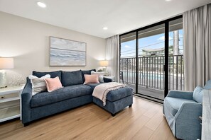 A modern living room with a sectional sofa, a small side table, a painting, and a view through a large sliding glass door. - Ocean Side Condo | Steps to the Beach (Ocean City)
