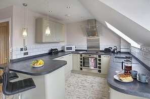 A modern kitchen with a built-in oven, microwave, and kettle. There is a fruit bowl on the counter and a coffee maker with cups and saucers. - 2 Bed in Seahouses (oc-cn166) (Seahouses)