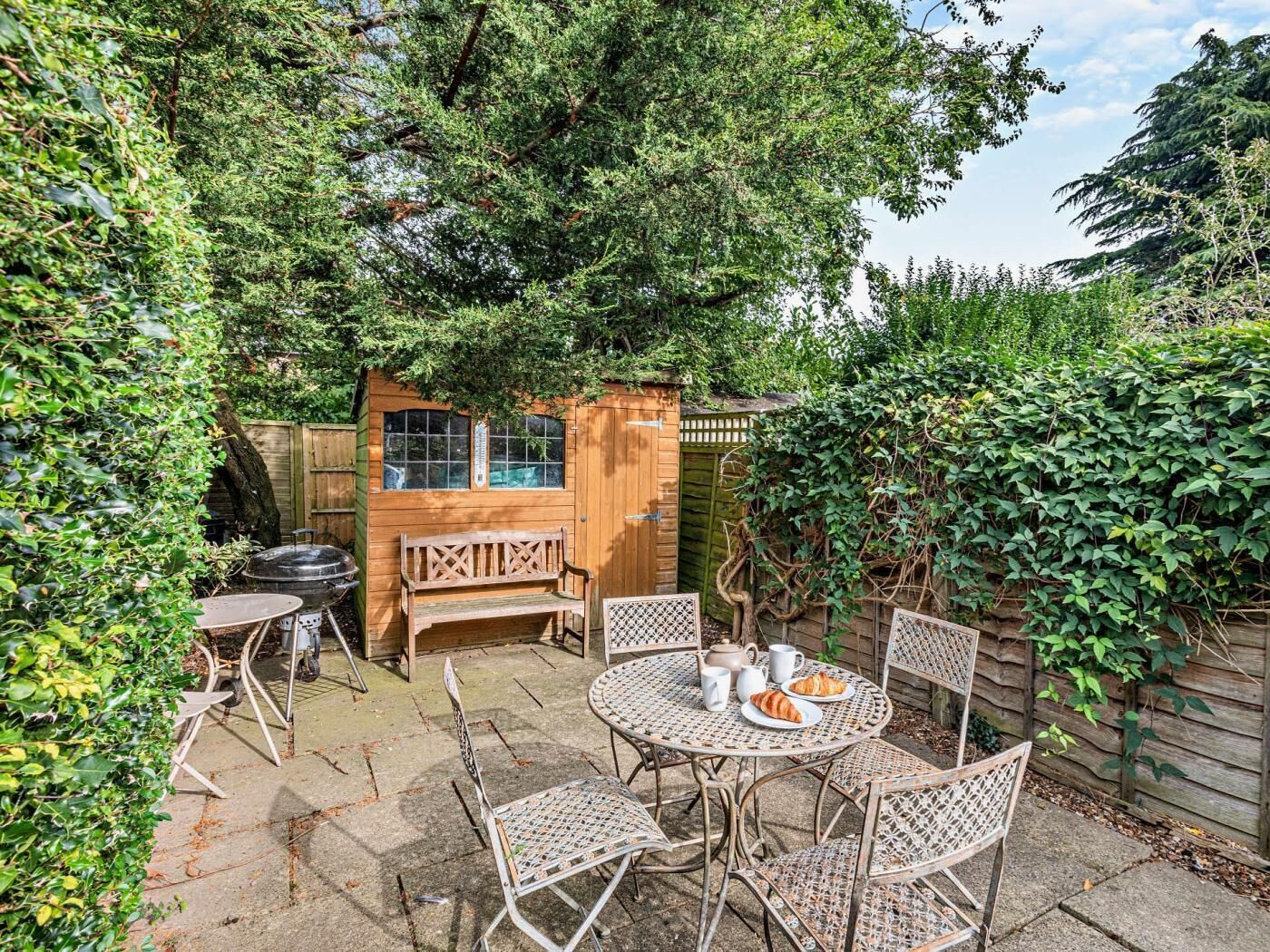 Een terras met een ronde tafel, vier stoelen, een houten bank en een klein houten bouwwerk met ramen.