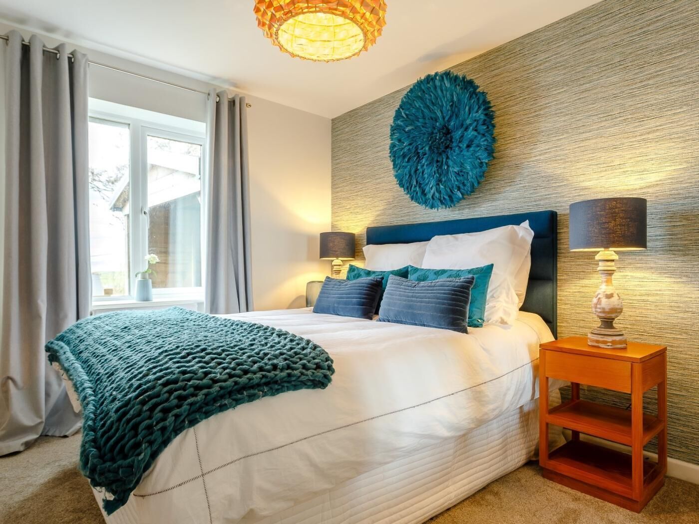 A bedroom with a bed, two bedside lamps, a wall-mounted decorative piece, and a window with curtains.