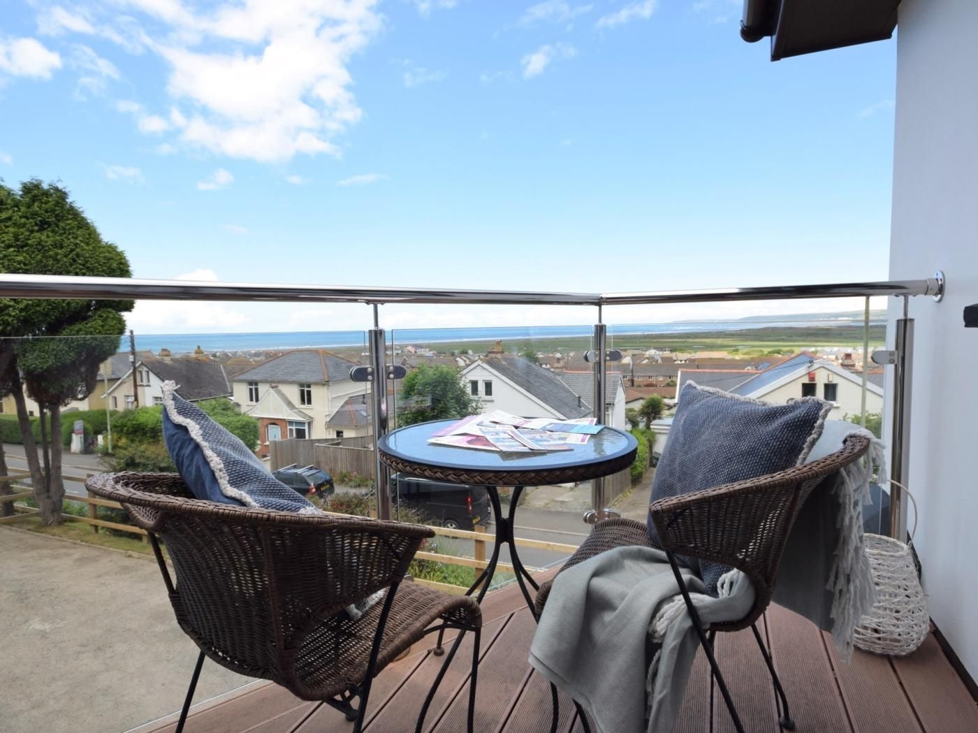 A balcony with wicker chairs and a small table, overlooking a residential area with houses and a view of the sea.