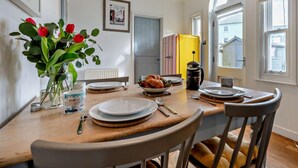 Ruokailutilassa on puinen pöytä, tuoleja, maljakko kukkineen, keltainen jääkaappi ja ikkuna, josta on näkymä rakennuksiin.