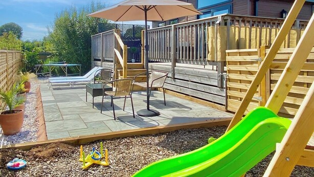 Un jardin avec un toboggan, une terrasse et un espace patio aménagé avec des chaises et un parasol.