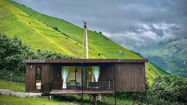 Una cabana de fusta amb un balcó, amb l'entorn de verdes muntanyes i un cel nuvolós.