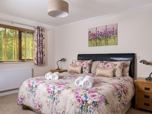 A bedroom with a bed, a nightstand, a lamp, a window with curtains, and a wall with a floral painting. - 4 Bed in Ystradgynlais (oc-bn343) (Cwm-twrch Isaf)