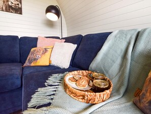 Un salon chaleureux avec un canapé bleu foncé, un panier en osier rempli de viennoiseries et une tasse de café, et une lampe fixée au mur.