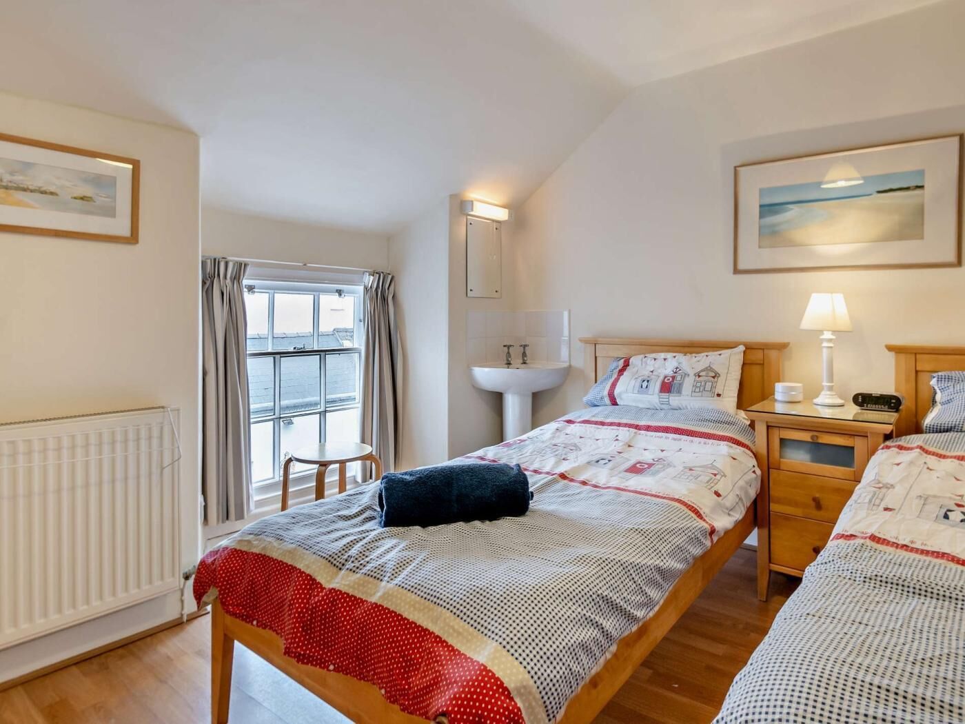 A bedroom with a bed, a bedside table with a lamp, a window with curtains, a radiator, and a wall-mounted light fixture.