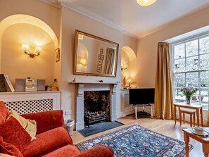 A living room with a fireplace, a television, a red sofa, and a large window with curtains. - 4 Bed in Tenby (oc-fb104) (Tenby)