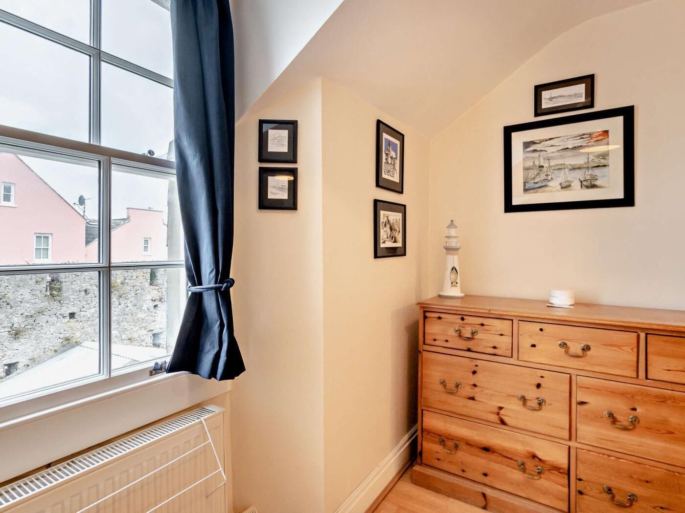 A room with a large window, a radiator, a wooden dresser, and several framed pictures on the wall.