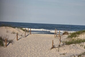 Ein sandiger Küstenpfad führt zum Meer, gesäumt von Holzbalken und Ketten.