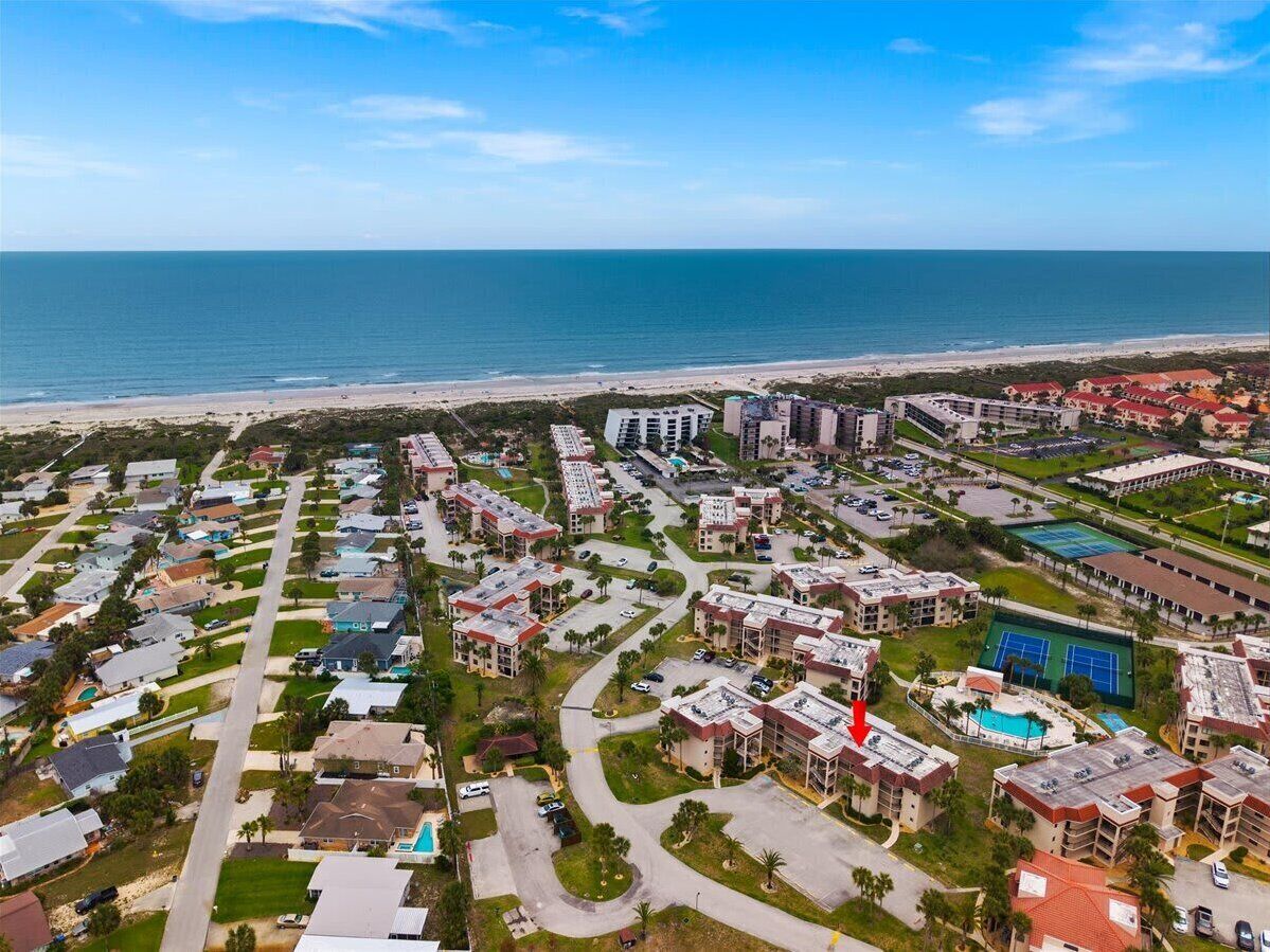 An aerial view of a coastal residential area with a beach, houses, and a large building with a pool.