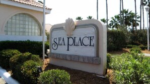 Un letrero que dice "SEA PLACE" con una concha decorativa en la parte superior, rodeado de arbustos bien cuidados y palmeras.