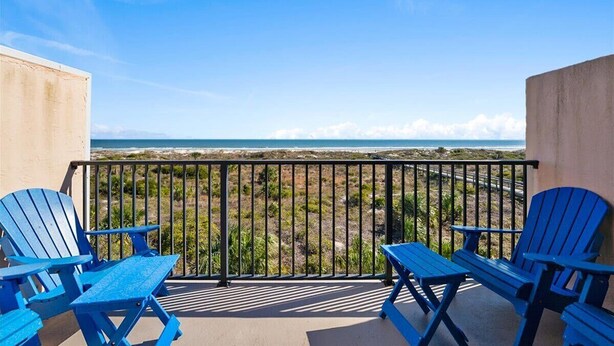 Un balcón con dos sillas Adirondack azules y una mesa, con vista a una playa y una vista despejada del océano.