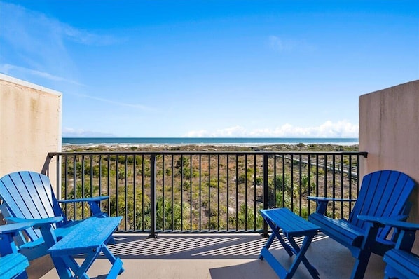 Ein Balkon mit zwei blauen Adirondack-Stühlen und einem Tisch, mit Blick auf einen Strand und eine klare Sicht auf den Ozean.
