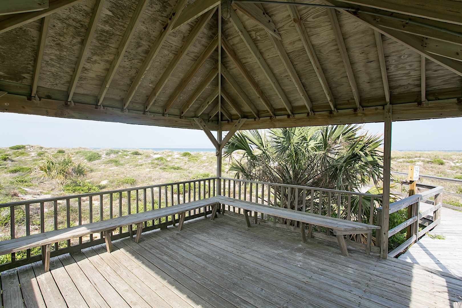 Ein hölzernes Pavillon mit einer Bank, der auf einen Strand mit spärlicher Vegetation und einen freien Blick auf den Ozean blickt.