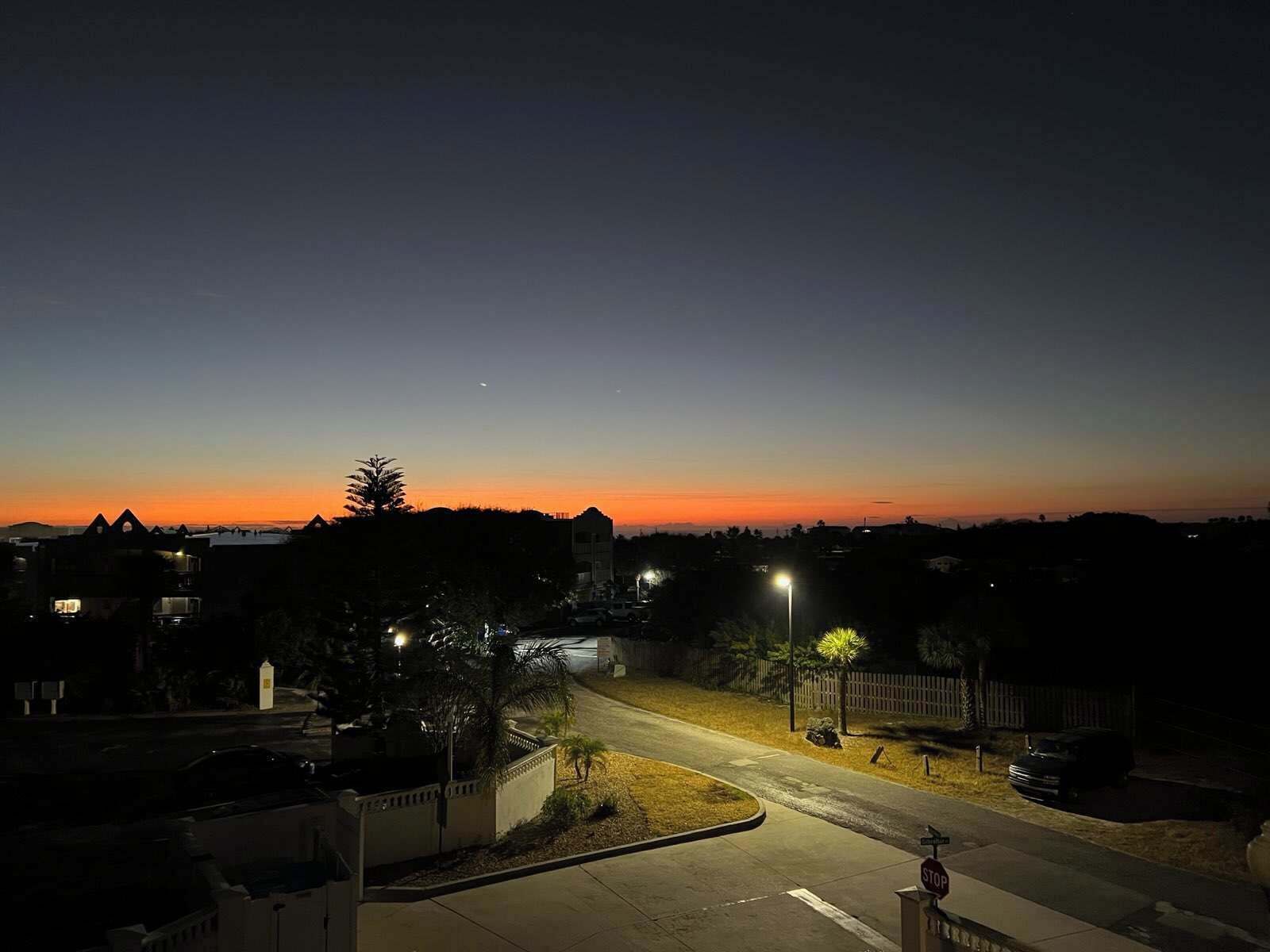 A residential street at dusk with houses, a stop sign, and streetlights.