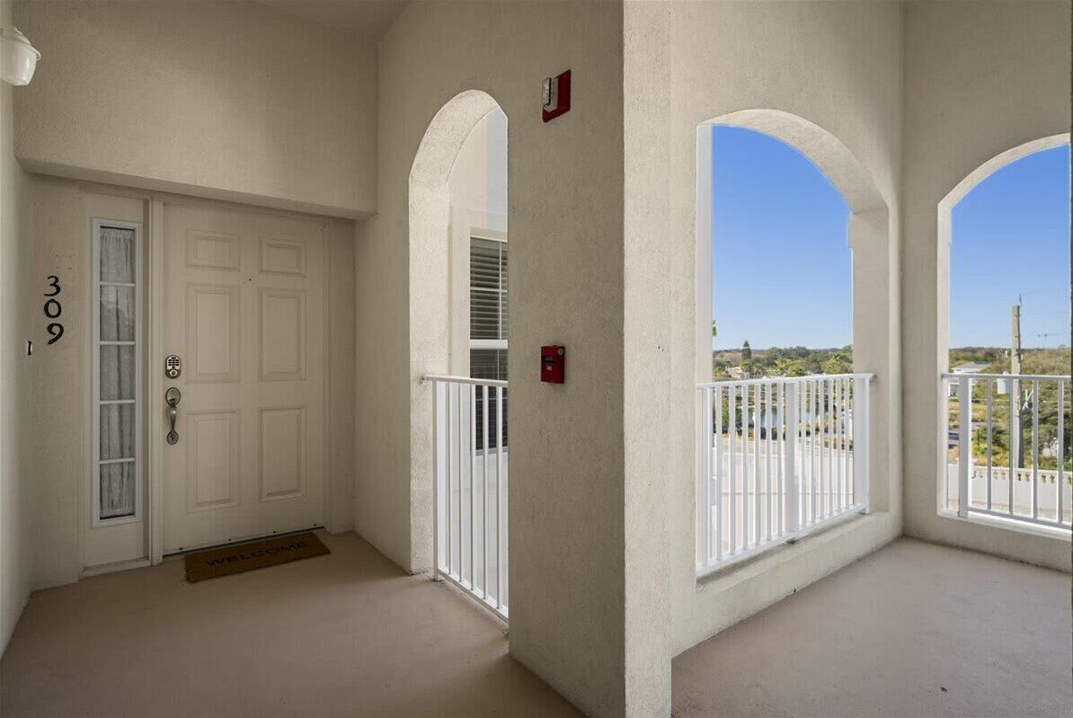 A clean, modern apartment entrance with a white door, a balcony with white railings, and a clear view of the surrounding area.