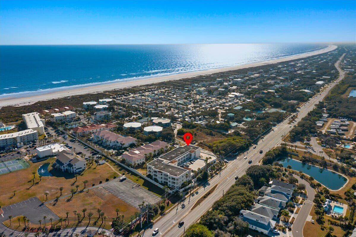 An aerial view of a coastal town with a prominent hotel marked by a red pin, surrounded by residential buildings, roads, and a beach.
