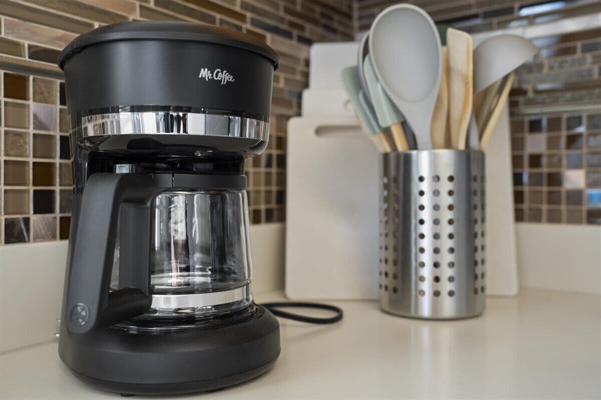A black coffee maker with a clear water reservoir and a metal drip tray, placed on a kitchen counter next to a stainless steel utensil holder with wooden spoons.