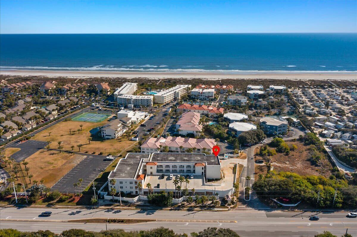 An aerial view of a coastal residential area with a large building complex, including a hotel with multiple floors and a parking lot.