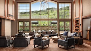 A spacious living room with large windows offering a view of a forested landscape. Furnishings include sofas, armchairs, and a coffee table.