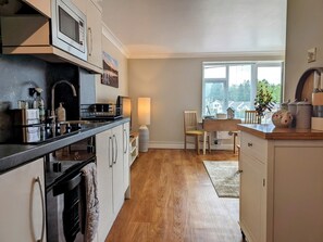 A modern kitchen with a central island, a dining area with a wooden table and chairs, and a view of the outside through large windows. - 1 Bed in Keswick (oc-sz468) (Keswick)