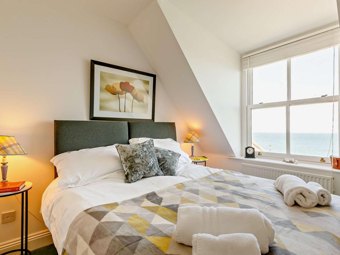 A hotel room with a bed, a framed picture on the wall, a window with a view, and a bedside table with a lamp.