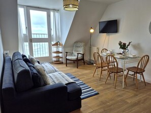 A modern living room with a sofa, dining table, chairs, a TV, and a large window. - 2 Bed in Ilfracombe (oc-87979) (Ilfracombe)