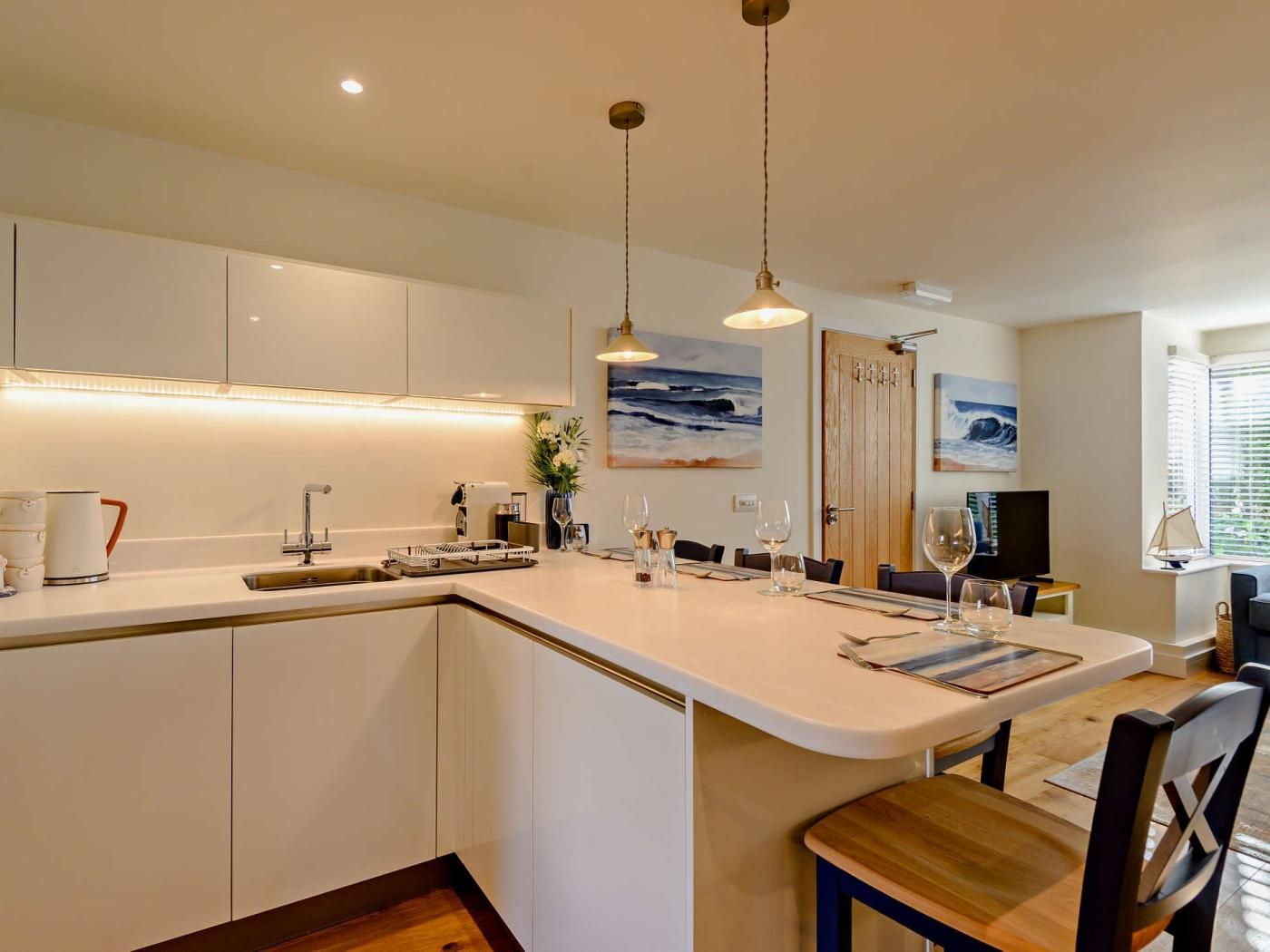 Une cuisine moderne avec des meubles blancs, un îlot central doté de sièges, des luminaires suspendus et des œuvres d’art accrochées aux murs.