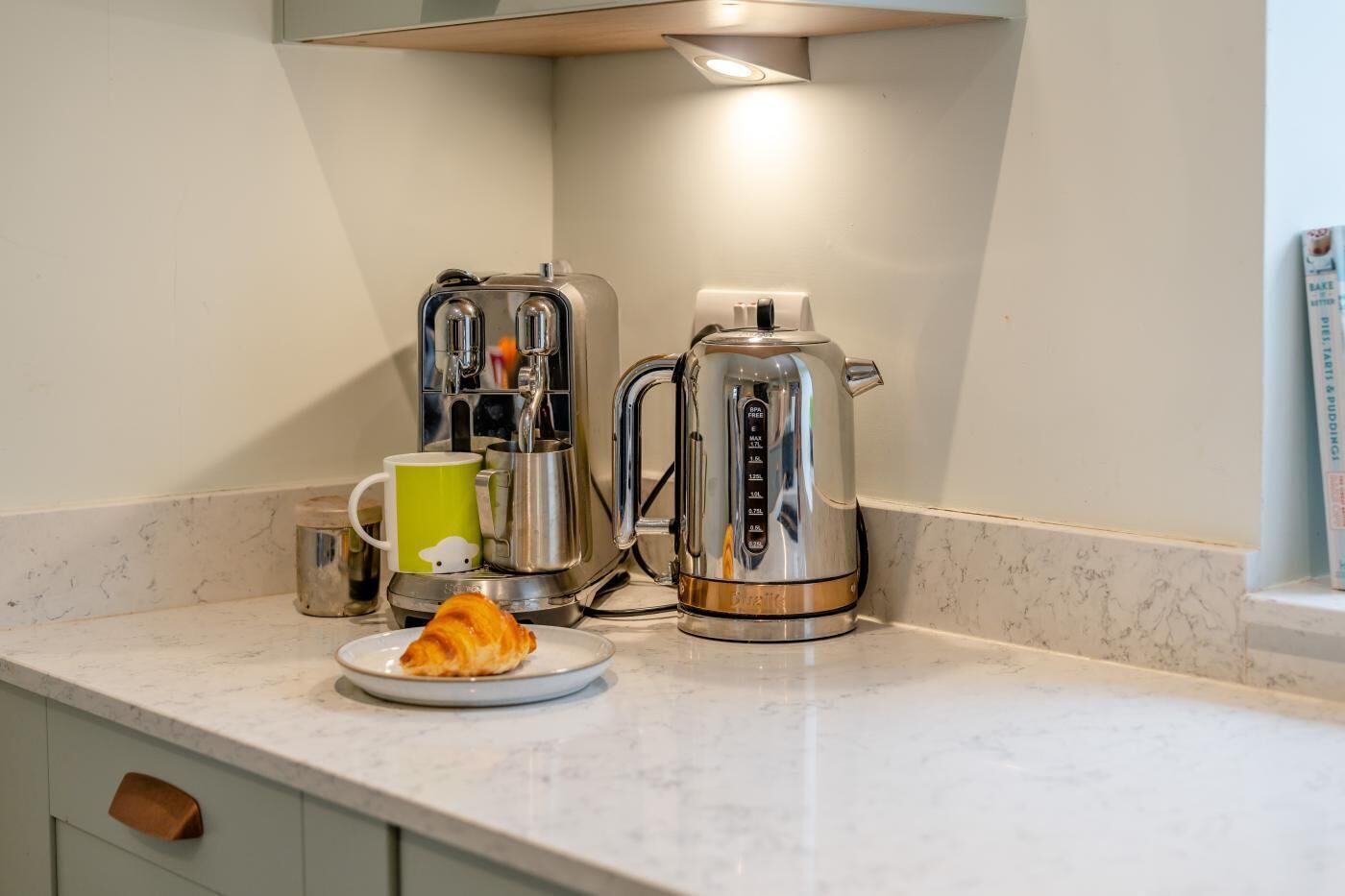 Een keuken met een koffiezetapparaat, waterkoker, mok en croissant op het aanrecht.