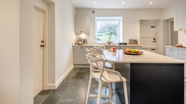 A modern kitchen with a large island, white cabinetry, and a window providing natural light. - 3 Bed in Broughton-in-furness (oc-91862) (Broughton Mills)