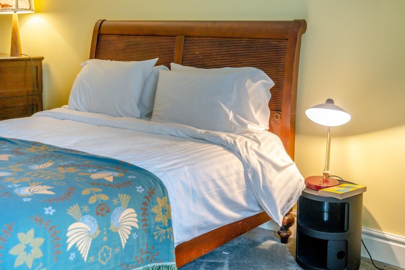 Una cama con ropa blanca de cama, una colcha floral azul, un cabecero de madera, una lámpara de noche y una mesita con un libro.