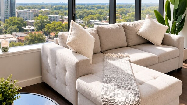 A modern living room with a large window, a beige sectional sofa, a glass coffee table, and a potted plant. - The Skyline Suite Midtown (Atlanta)