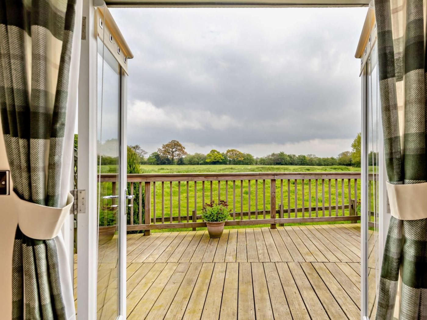 A wooden deck with a view of a grassy field and trees, a wooden railing, and a potted plant.
