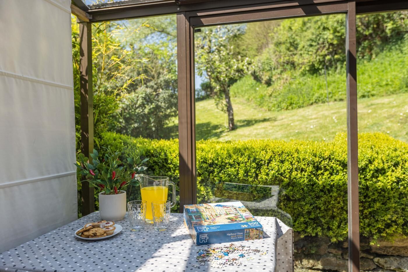 Una mesa con una caja de acertijos, un plato de galletas y vasos de jugo de naranja en un espacio exterior cubierto con vistas a un jardín.
