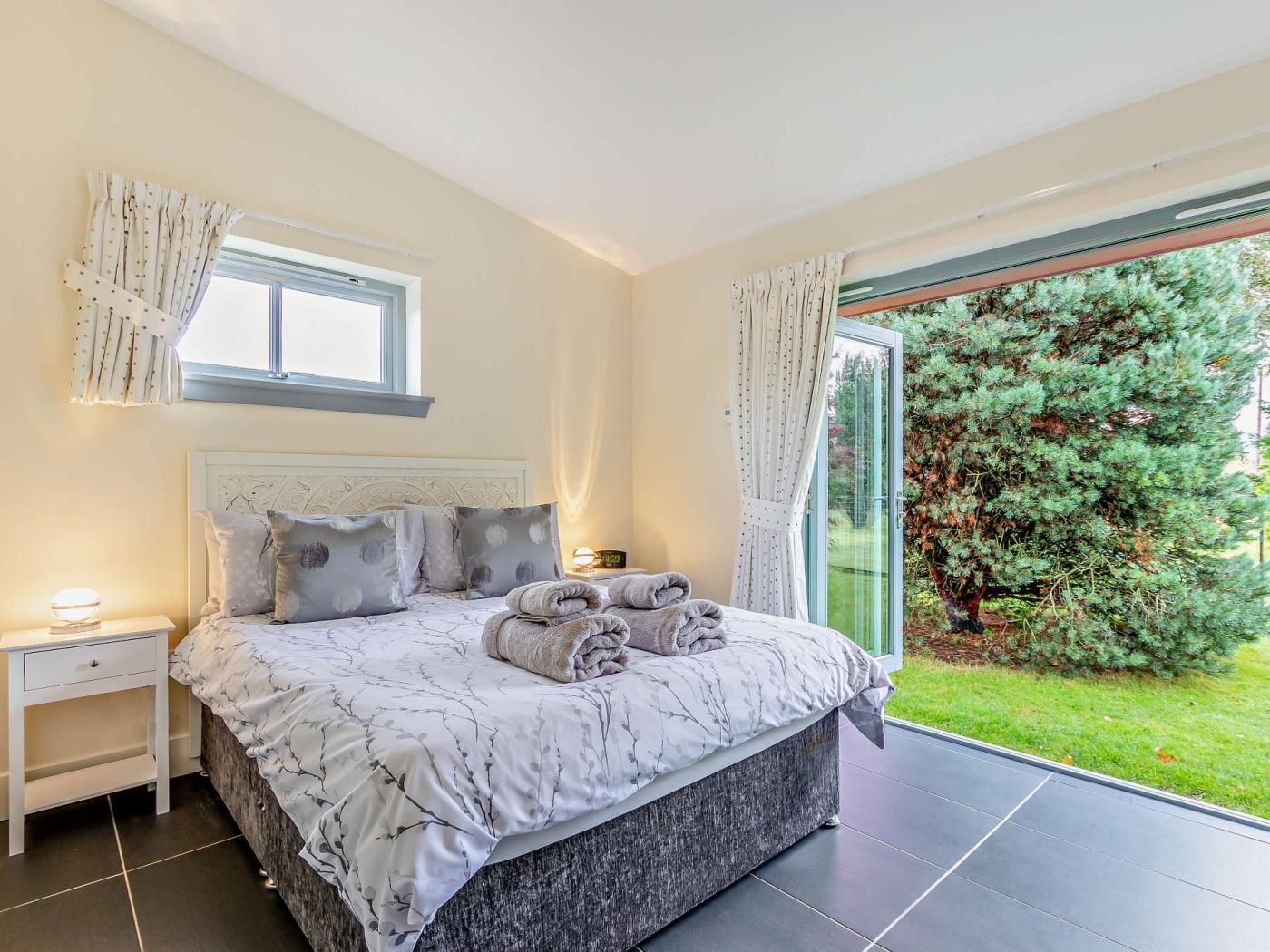 A bedroom with a bed, nightstands, a window, and a view of a garden.