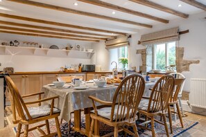 Un coin repas chaleureux avec des chaises en bois et une table dressée pour quatre personnes, agrémenté d’une cuisine rustique en arrière-plan, avec un mur en pierre et une fenêtre offrant une vue agréable.