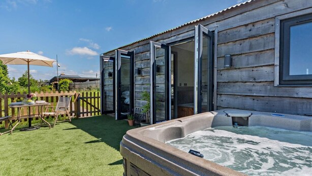 A wooden cabin with a hot tub, a patio with a table and chairs, and a wooden fence.