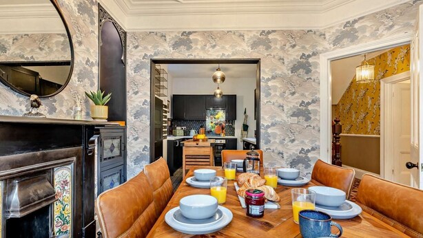 Un coin repas avec une table en bois dressée pour le petit-déjeuner, une cheminée et une cuisine en arrière-plan.