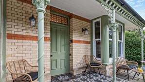 Un bâtiment en briques avec une porte verte, deux chaises en osier et une terrasse carrelée.