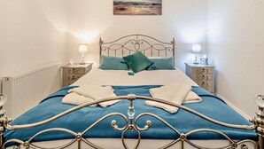 Ein ordentlich bezogenes Bett mit blauer Bettdecke und zwei Kissen, begleitet von Nachttischen mit Lampen und einem Wandbild darüber.