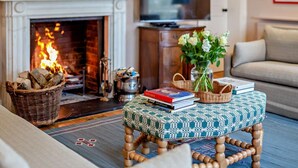 A cozy living room with a fireplace, a television, a sofa, a coffee table with books, and a vase with flowers. - 8 Bed in Dorchester (oc-93723) (Minterne Magna)